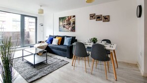 Un salon moderne avec un canapé foncé, une table basse en verre et un coin repas comprenant une table blanche et des chaises grises.