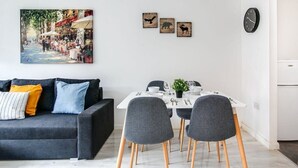 Un salon moderne avec un canapé gris foncé, une table à manger blanche et quatre chaises grises, ainsi qu’une petite plante en pot.