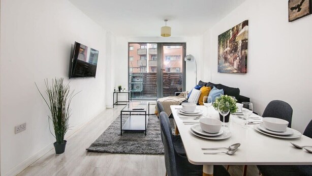 Un salon moderne comprenant un coin repas, une plante en pot, un téléviseur à écran plat et une grande fenêtre donnant sur des immeubles.