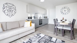 Ein modernes Wohnzimmer mit Sofa, Esstisch mit Stühlen, Küchenzeile und Wanduhr.