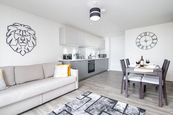 A modern living room with a sofa, a dining table with chairs, a kitchen area, and a wall clock. - Manhattan View | Netflix | Fast WiFi (West Midlands)
