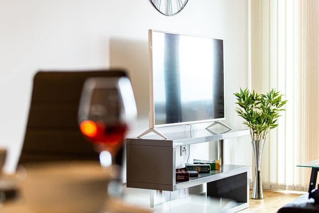 Ein modernes Wohnzimmer mit Flachbildfernseher an der Wand, einem Glastisch, einem dunkelholzigen Beistelltisch, einer Vase mit Grünpflanzen und einer Wanduhr.