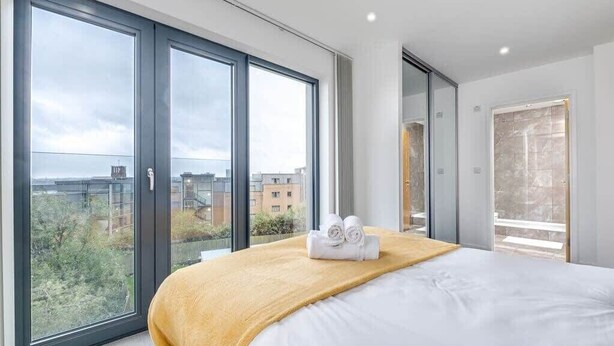 Habitación de hotel moderna con una cama grande, una manta amarilla y vistas de la ciudad a través de ventanales del piso al techo.