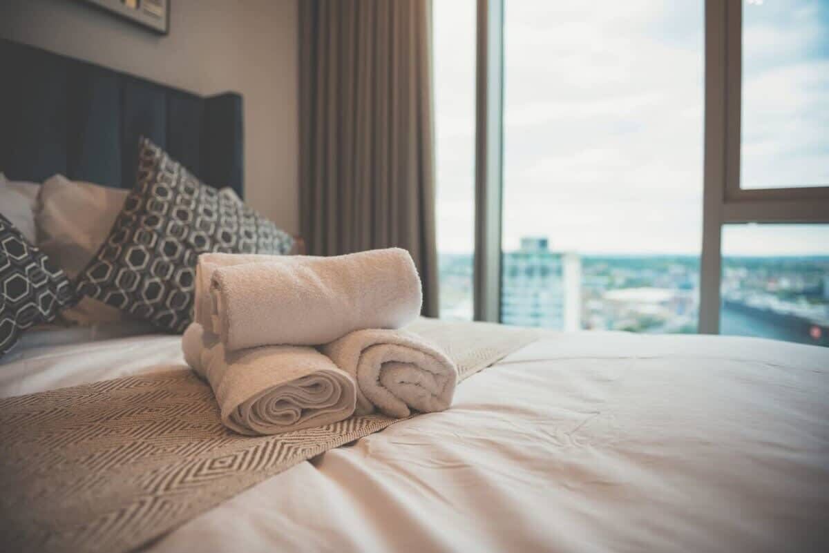 A neatly made bed with rolled towels and patterned bedding in a hotel room with a large window offering a city view.