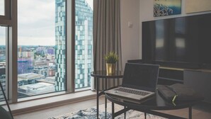 A modern living room with a large window, a laptop on a small table, a sofa, and a TV.