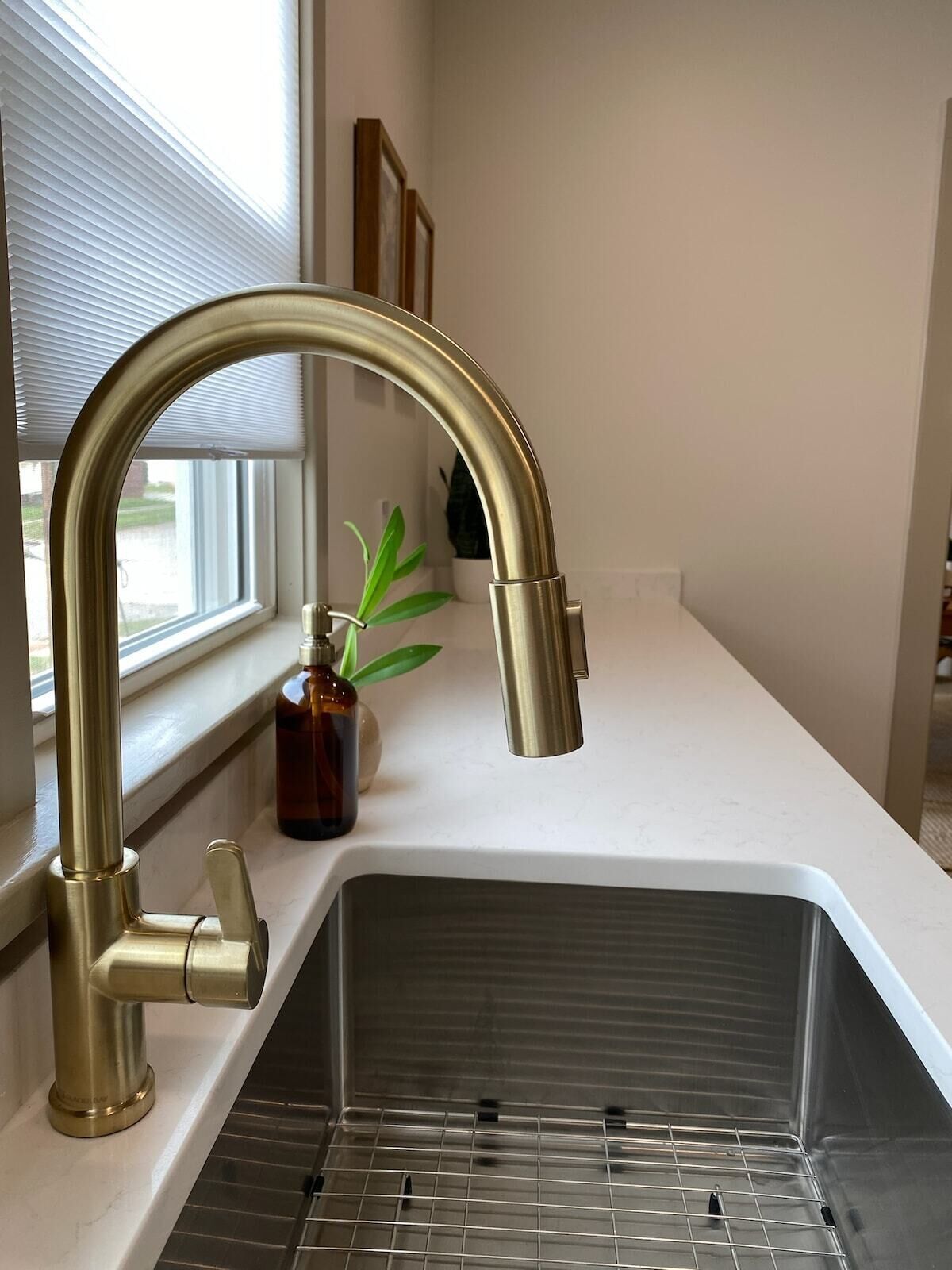 Eine Küche mit einer Spüle aus Edelstahl, einem goldfarbenen Wasserhahn und einem Fenster mit Jalousien.