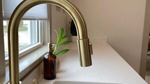 Una cocina con un lavamanos de acero inoxidable, un grifo color dorado y una ventana con persianas.