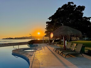 A pool area with lounge chairs and a thatched umbrella, overlooking a body of water at sunset. - Villa Familiar Frente al mar (Cruz de Huanacaxtle)