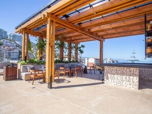 Relax on the rooftop terrace with a sofa set, chairs, and a bar area, surrounded by potted plants and an open view of the city.