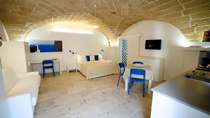 Quarto compacto com cama, escrivaninha e área de refeições, apresentando teto de pedra e paredes brancas.