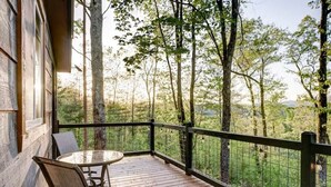 A wooden deck with a glass table and chairs, surrounded by a metal railing, overlooking a forested area.