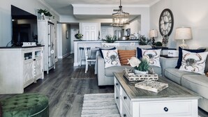 Ein modernes Wohnzimmer mit einem grauen Sofa, einem Couchtisch und einem grünen Ottomanen. Im Hintergrund ist eine Küche mit weißen Schränken und einem Essbereich zu sehen.