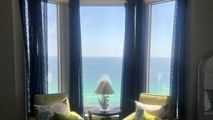Ein Zimmer mit großem Fenster, zwei Sesseln, einem kleinen Tisch mit Lampe und einem Bett mit Blumenmuster-Überwurf.