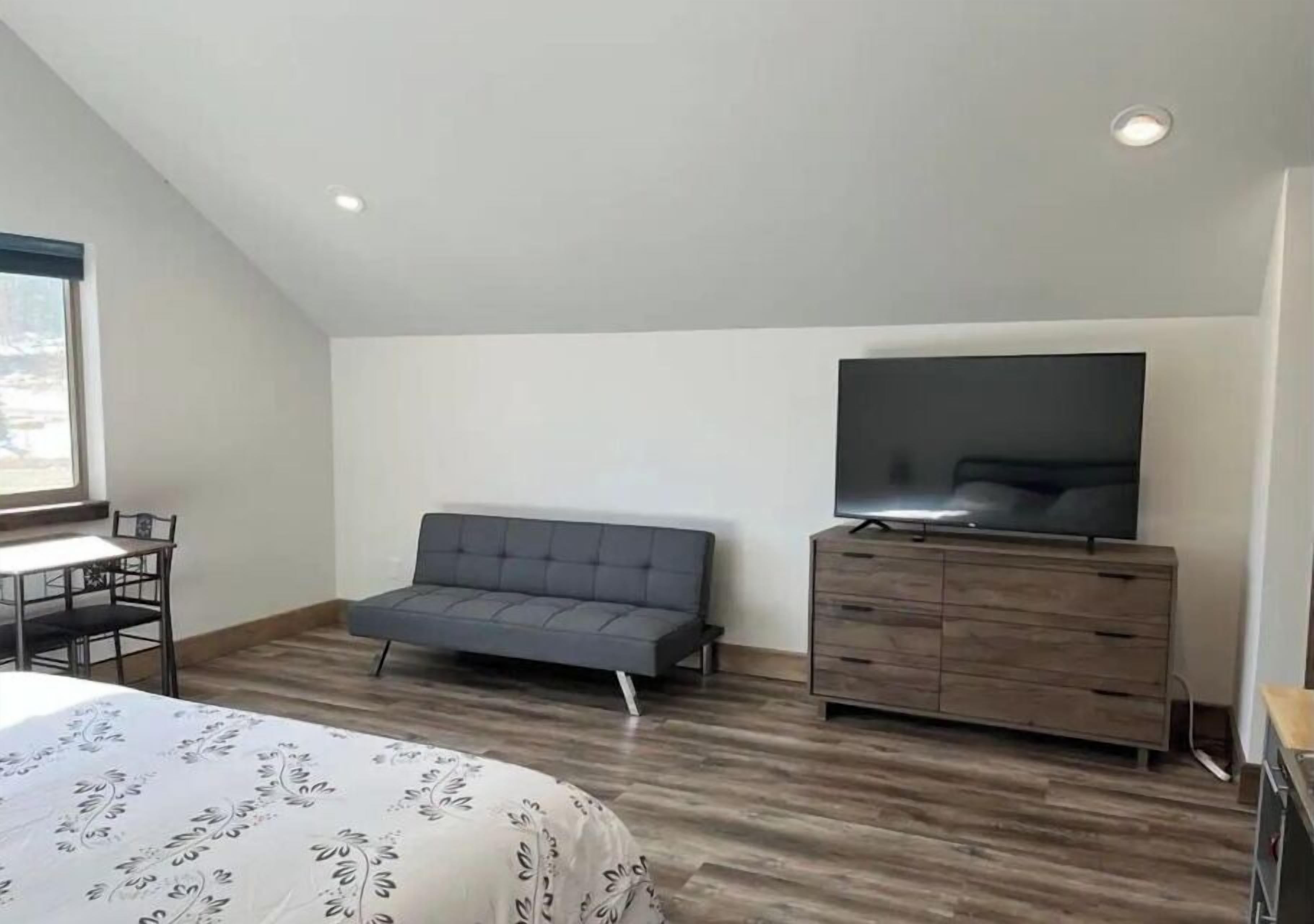 A bedroom with a bed, a grey sofa, a TV on a wooden cabinet, and a window.