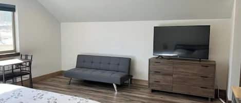 A bedroom with a bed, a grey sofa, a TV on a wooden cabinet, and a window.