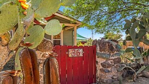 Un bâtiment rustique avec une porte rouge, entouré de cactus et d’un mur de pierre.