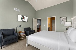 Una habitación de hotel con una cama grande, dos sillones, una mesita de noche con lámpara, un cuadro enmarco y una puerta que da al baño.