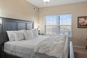 A bedroom with a large bed, a nightstand with a lamp, a window with blinds, and a painting on the wall.