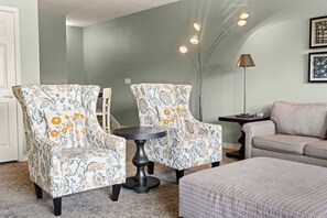 Una sala de estar con dos sillones estampados, una mesa de centro redonda, un sofá, una lámpara de pie y una obra de arte abstracta enmarcada en la pared.