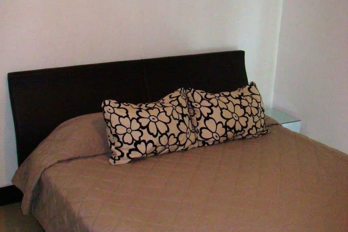 A bed with a patterned pillow and a plain headboard in a room with plain walls.