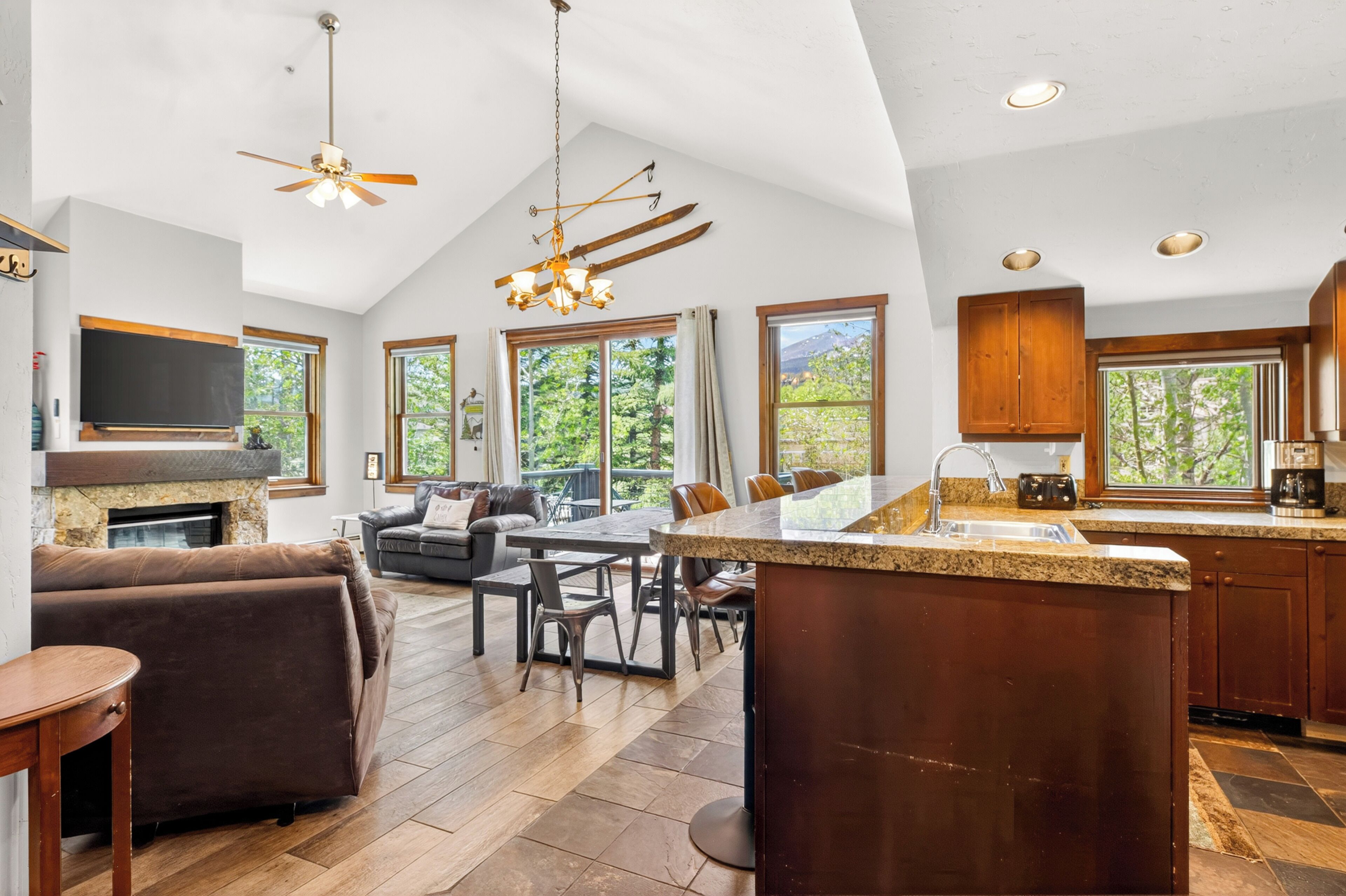 A spacious kitchen with a large island, a dining area with a table and chairs, a fireplace, and large windows offering a view of greenery.