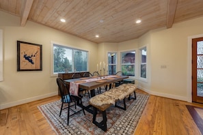Une salle à manger avec une table en bois, des chaises et un banc. Il y a des fenêtres donnant sur des arbres et une porte avec un panneau de verre.