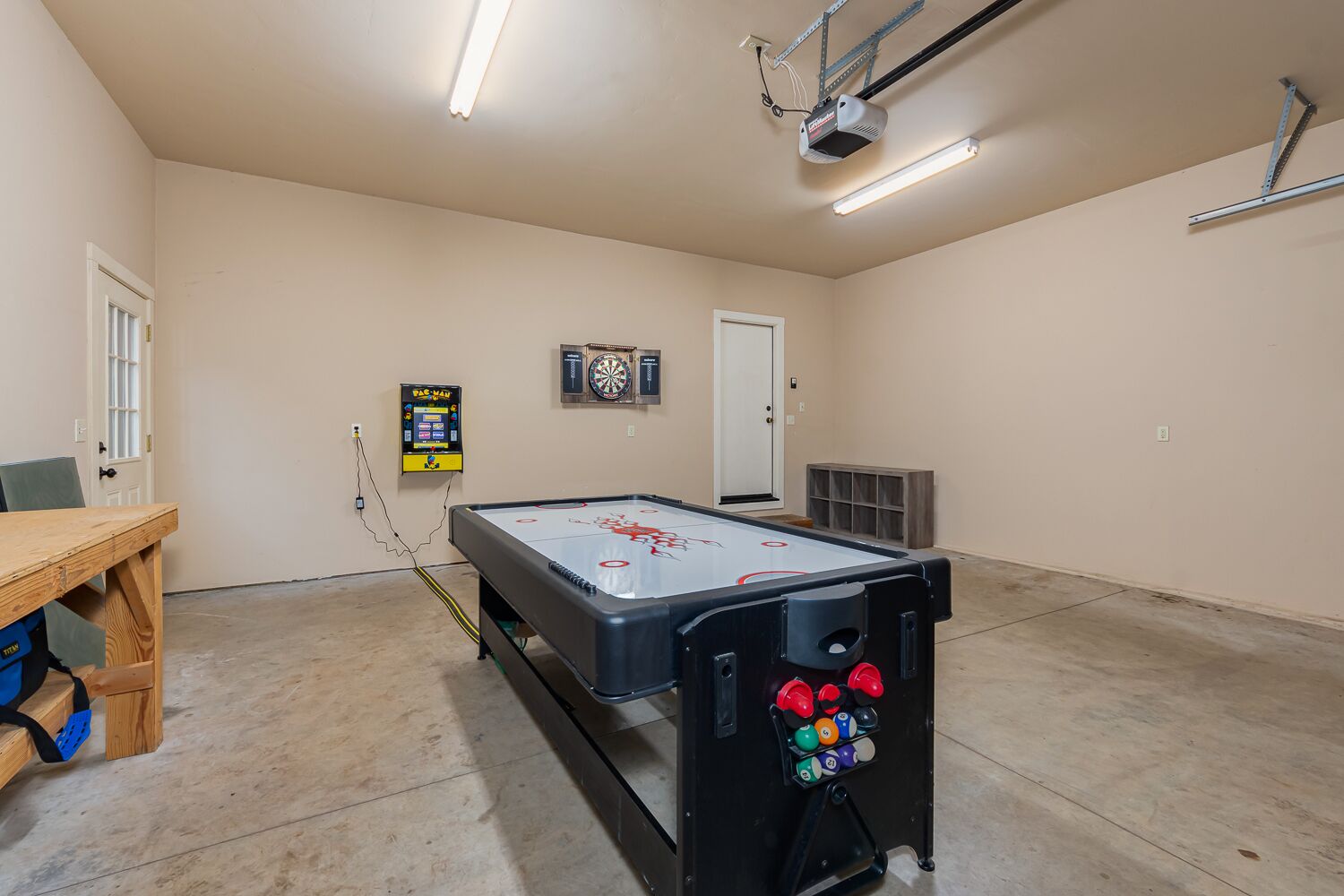 Ett biljardbord med en darttavla och en Pac-Man-spelautomat i ett rum med beige väggar och en dörr.