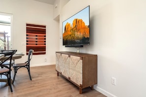 A modern living room with a wooden dining table, chairs, a flat-screen TV mounted on the wall, and a decorative wall panel.