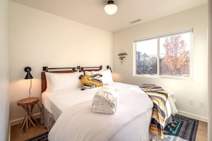 A bedroom with a bed, a bedside table, a lamp, a window with a view of trees, and a wall-mounted light fixture.