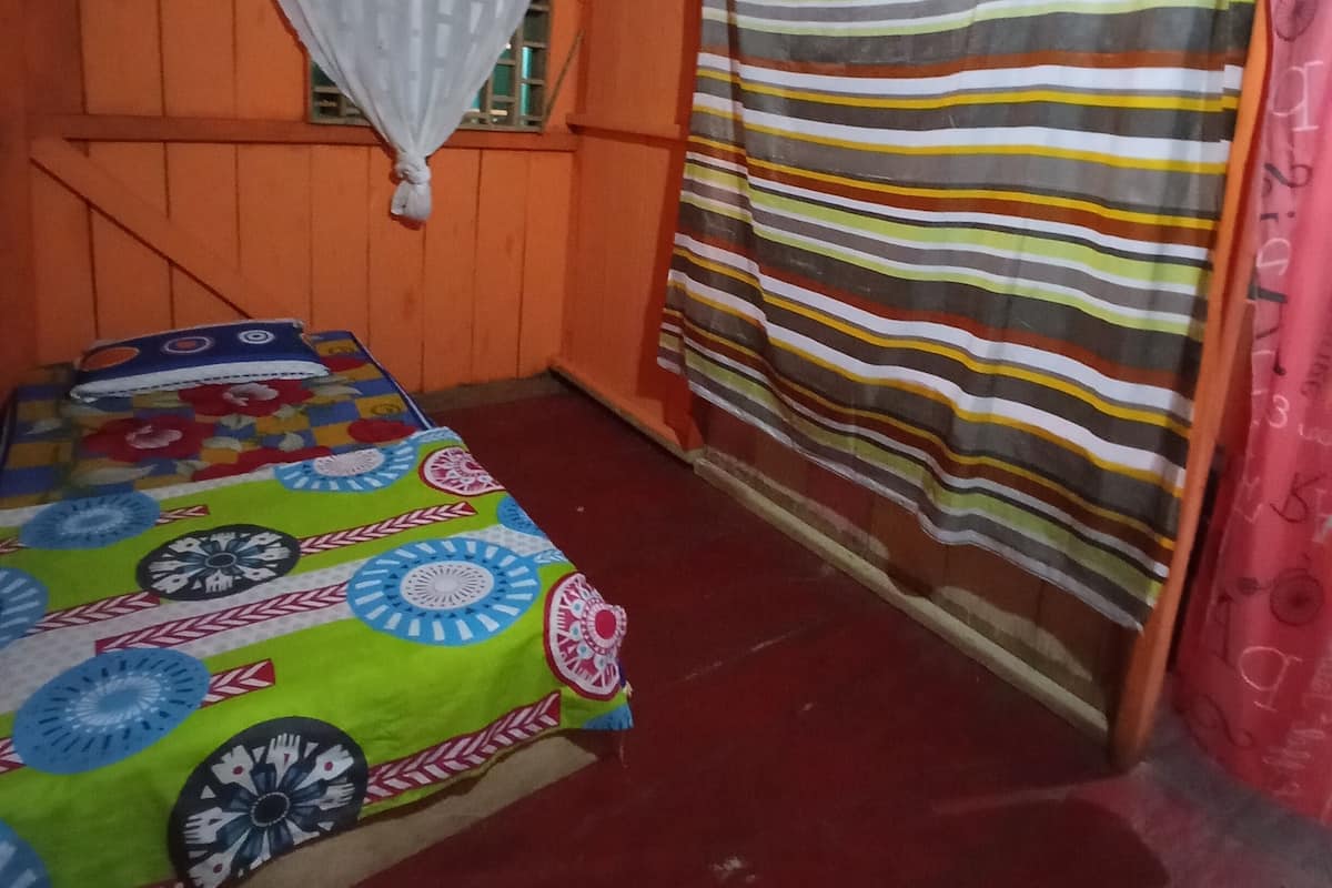 A room with a bed covered in a colorful floral bedspread, an orange wall, a striped curtain, and a white curtain tied at the top.