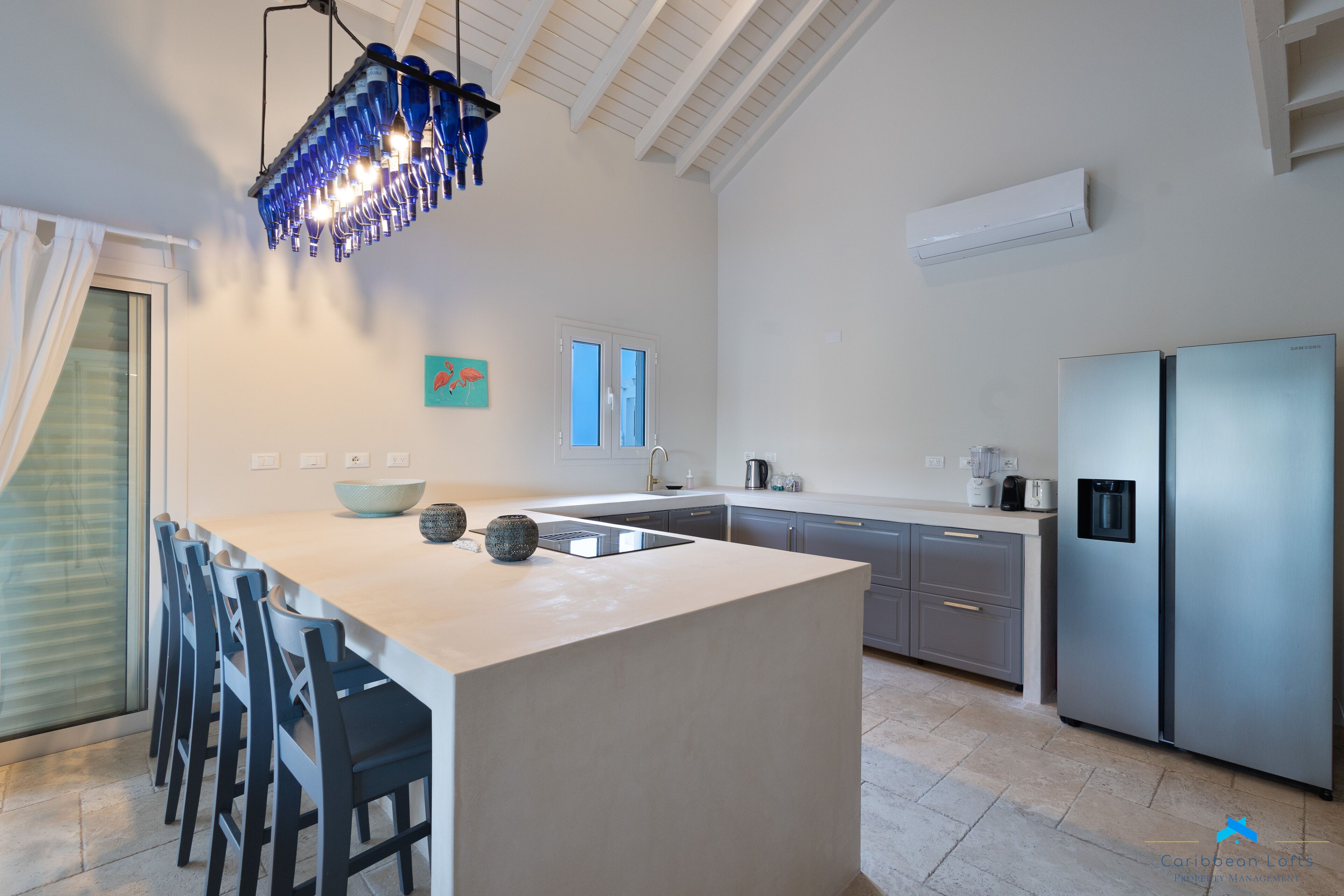 A modern kitchen with a central island, bar stools, a stainless steel refrigerator, and a unique blue pendant light.