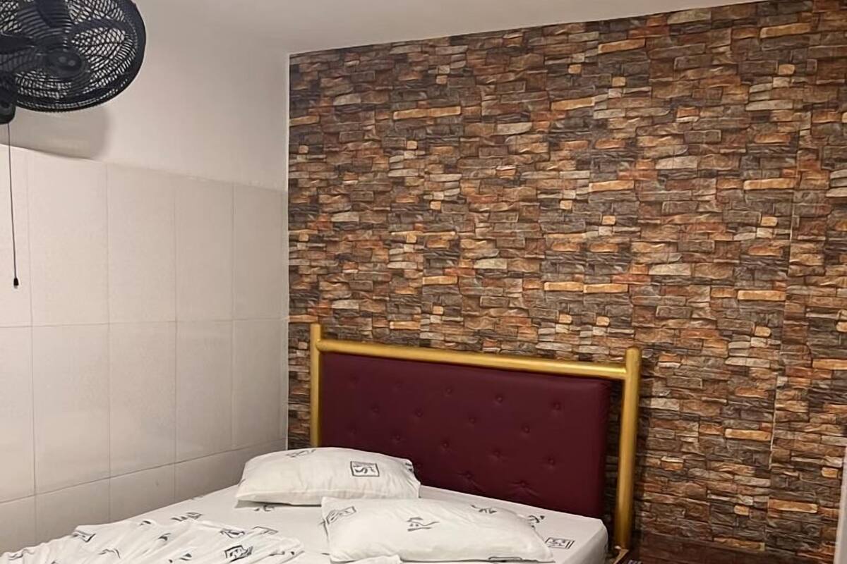 Una cama bien hecha con ropa blanca y un cabecero rojo, una mesita de noche de madera y una pared con textura similar a la piedra.