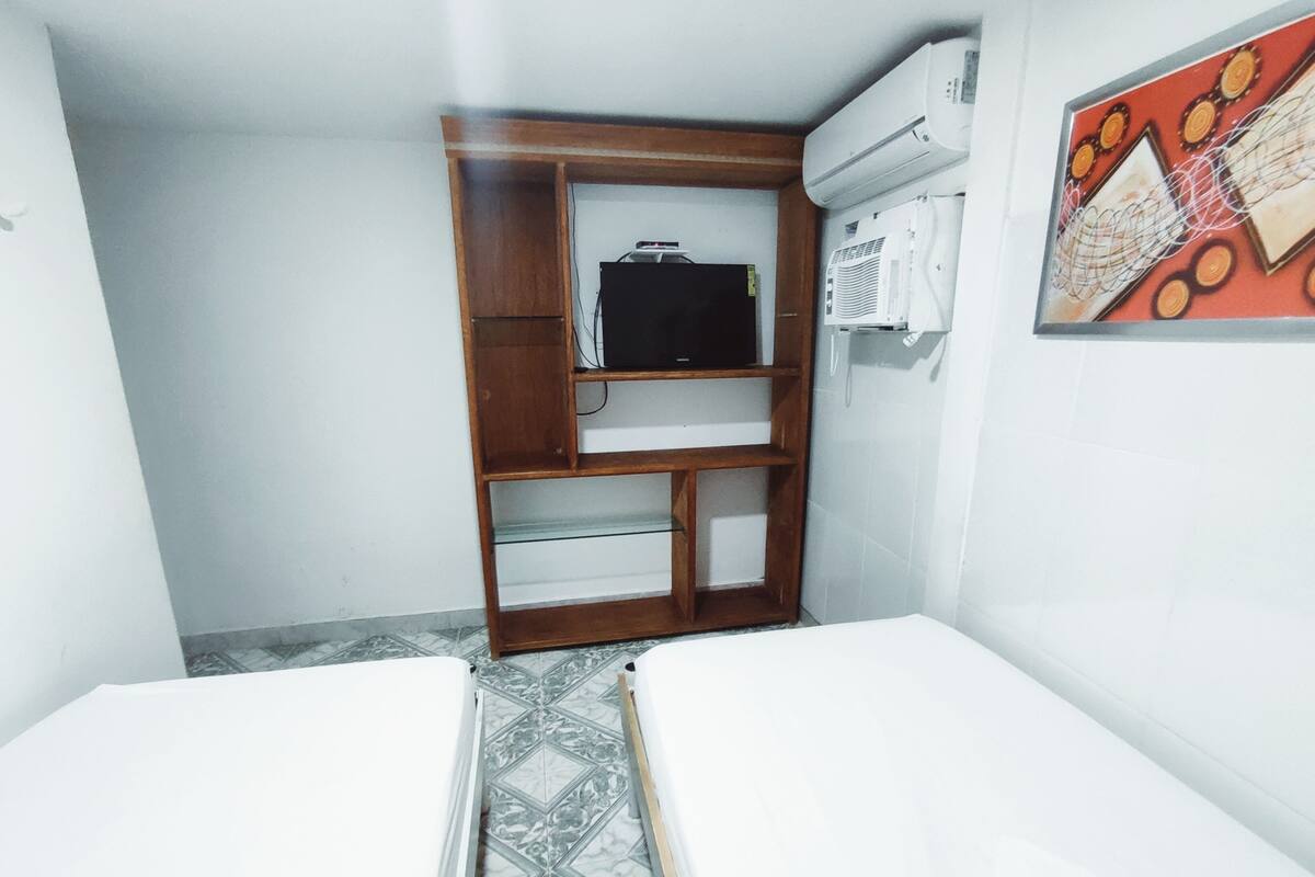 Una habitación pequeña y funcional con una cama, un armario de madera con televisor y un equipo de aire acondicionado.