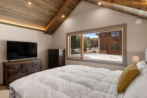 A bedroom with a large bed, a television on a wooden dresser, and a window with a view of a snowy landscape. - Deluxe Ski-In/Out Home! Walking Distance to Resort ! Cozy Fireplace! (Big Sky)