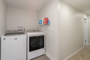 A laundry room with two washers and a dryer, a shelf with detergent, and a door leading to another room.