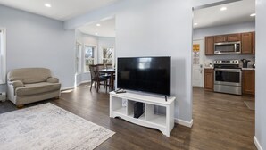 Un salon moderne avec un téléviseur à écran plat posé sur un meuble blanc, un canapé beige et un coin repas comprenant une table et des chaises en bois.