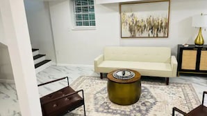 Una sala de estar con un sofá blanco, una mesa de centro redonda, un tapete estampado, un cuadro en la pared y una lámpara sobre una mesita auxiliar.