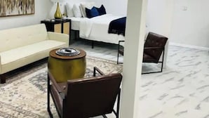 Una sala de estar moderna con un sofá blanco, una mesa de centro redonda, una cama con ropa de cama blanca y almohadas azules, y un cuadro grande en la pared.