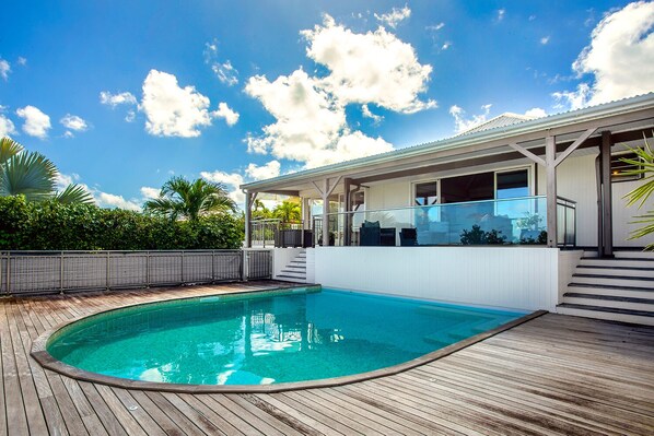 Piscina com deck de madeira, área de varanda coberta e vista para uma casa com fachada branca e varanda.