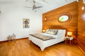 Quarto com cabeceira de madeira, um claraboia redonda, pintura de Buda, cama com roupa de cama branca, duas luminárias de cabeceira e duas gaiolas de pássaros.