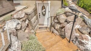 A stone staircase leading to a door with a decorative glass panel, surrounded by large rocks and a small garden area. - The Suite at the Cedars (Cedar Hills)