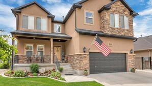 A two-story house with a stone facade, a front porch with outdoor seating, and an American flag on the garage door. - The Suite at the Cedars (Cedar Hills)