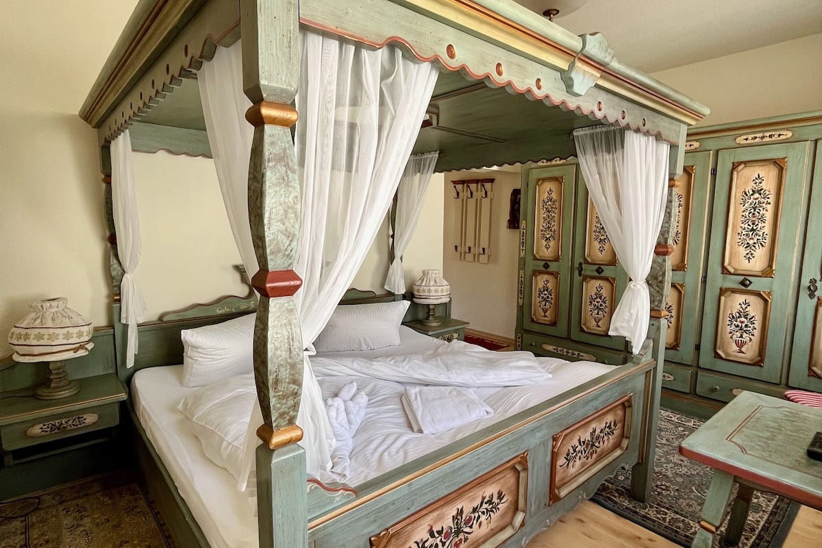 Una cama con dosel con ropa de cama blanca y un dosel, una mesita de noche de madera con una lámpara, y una pequeña mesa con una silla.