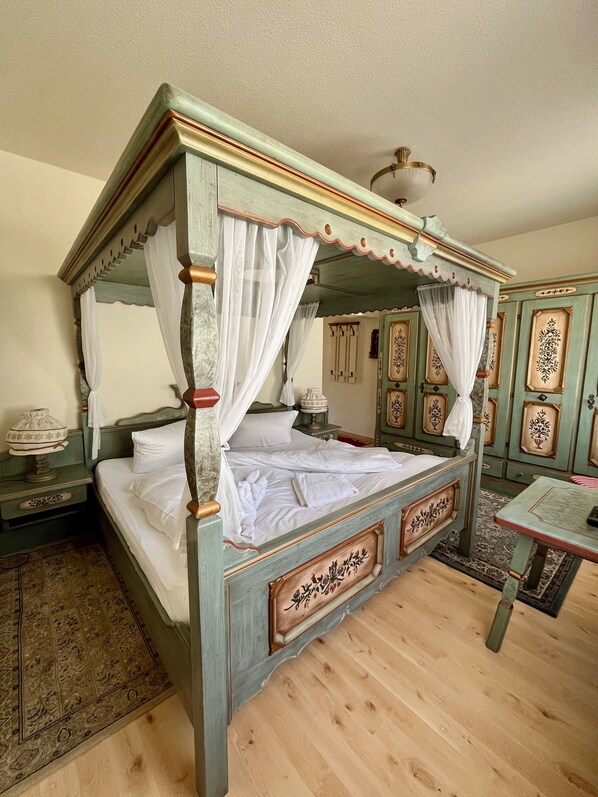 A four-poster bed with white linens and a canopy, a wooden nightstand with a lamp, and a small table with a chair. - Landhaus-Chalet-Keilberger Blick  (Oberwiesenthal)