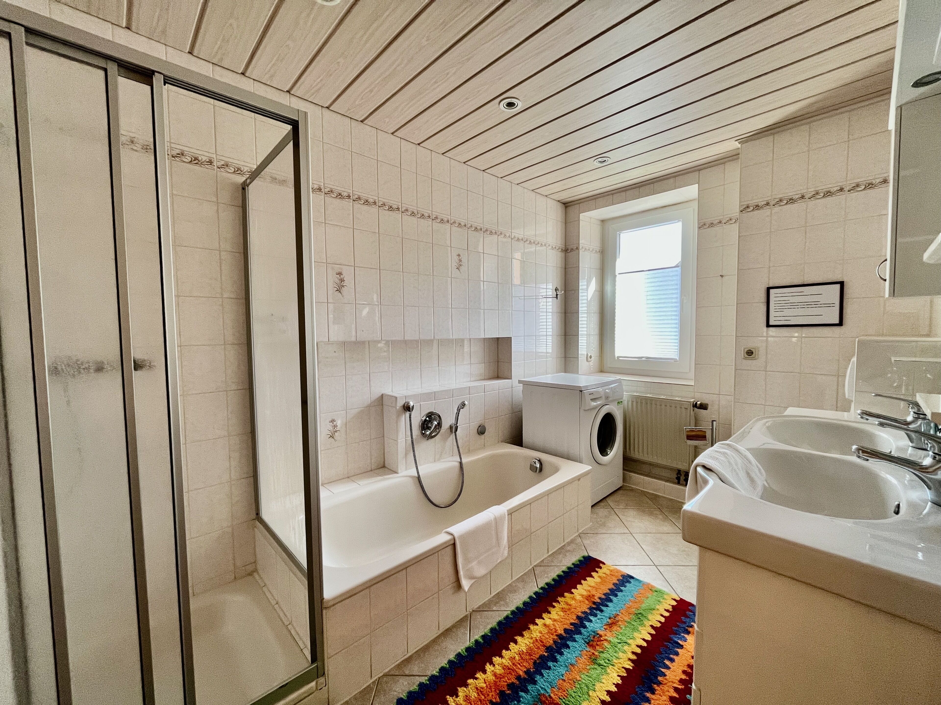 A bathroom with a glass shower enclosure, a bathtub, a washing machine, and a sink.