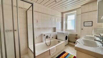 A bathroom with a glass shower enclosure, a bathtub, a washing machine, and a sink.