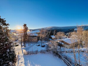 Schneebedeckte Landschaft mit einem Gebäude, das einen markanten Schornstein hat, umgeben von Bäumen und einem klaren Himmel.