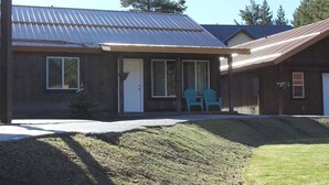 A two-story building with a metal roof, a front porch with two blue chairs, and a small garden area.