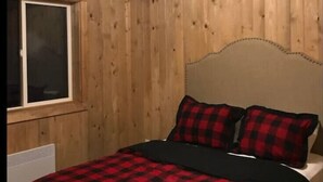 A room with wooden paneled walls, a bed with a red and black checkered bedspread, and a window with blinds.