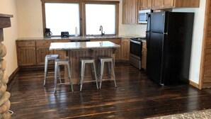 Une cuisine avec des meubles de rangement en bois, un réfrigérateur noir, un îlot central avec des tabourets et un lustre.